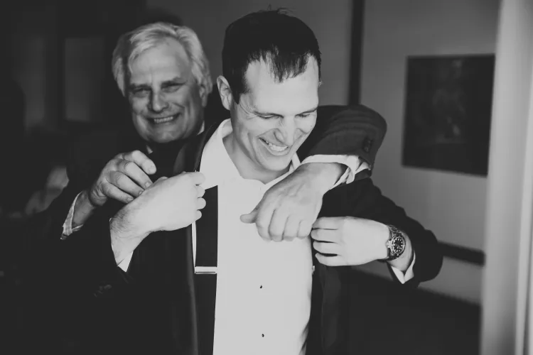 A groom laughing with an older man who helps him into his jacket, in black and white