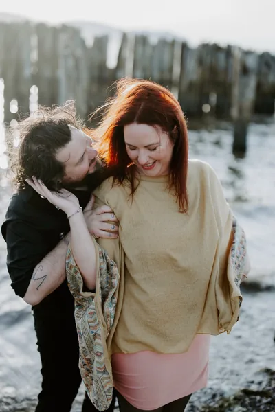 A woman laughing as her partner leans in, beside a wooden pier
