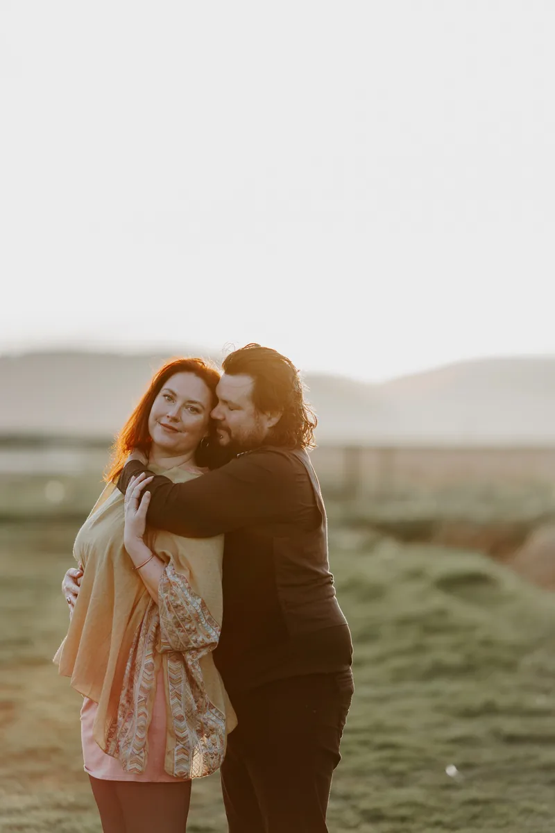 A couple holding each other in a meadow, lit from behind by a low sun